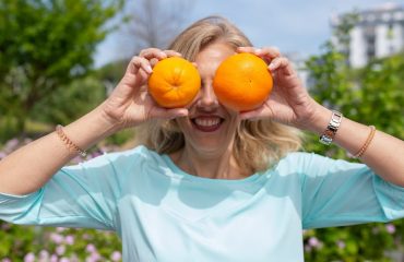 A smiling woman playfully holding oranges in a sunny park setting.